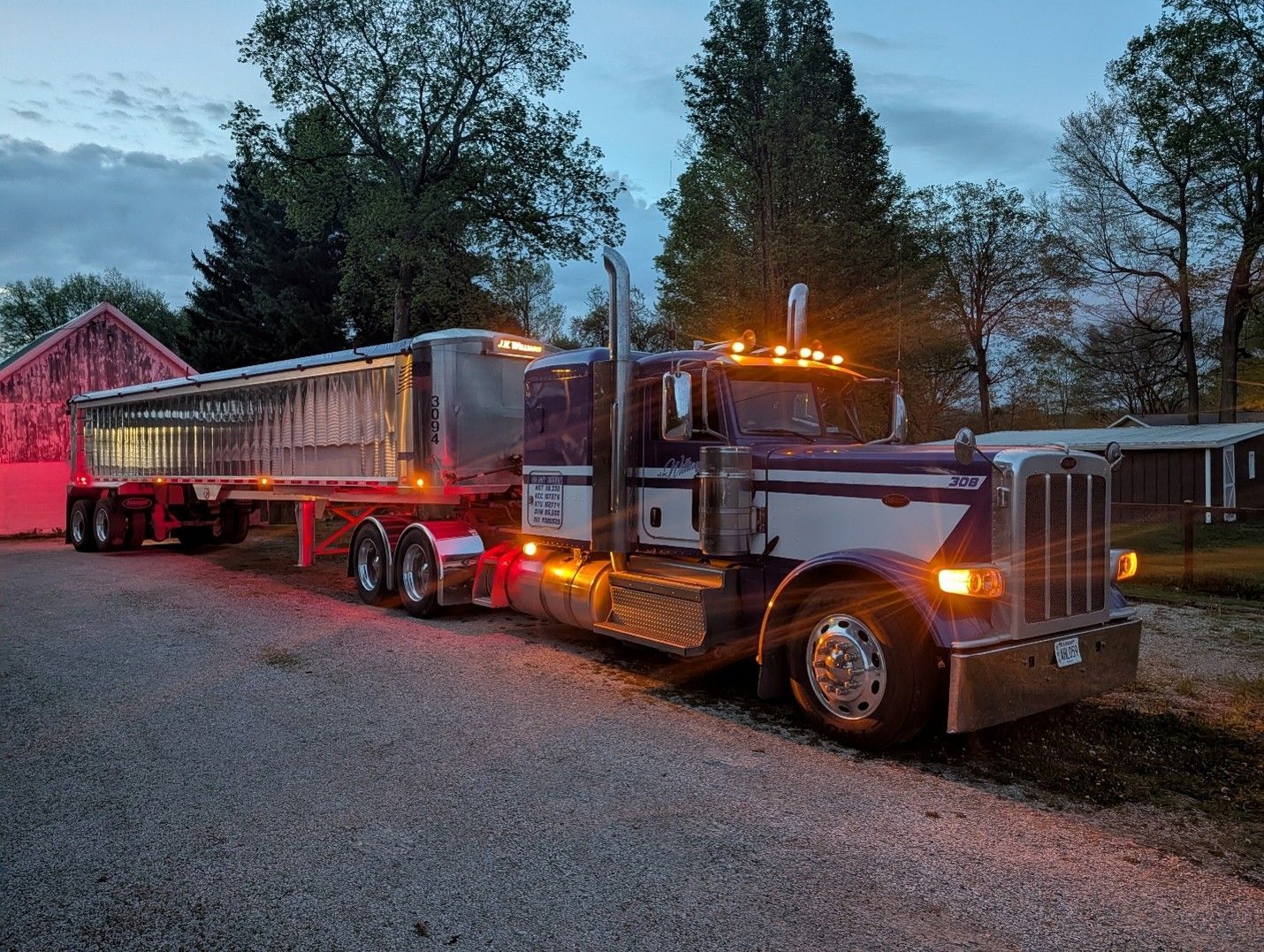 Silver semi-truck with trailer parked on a gravel lot at dusk near trees and a barn, lights glowing.
