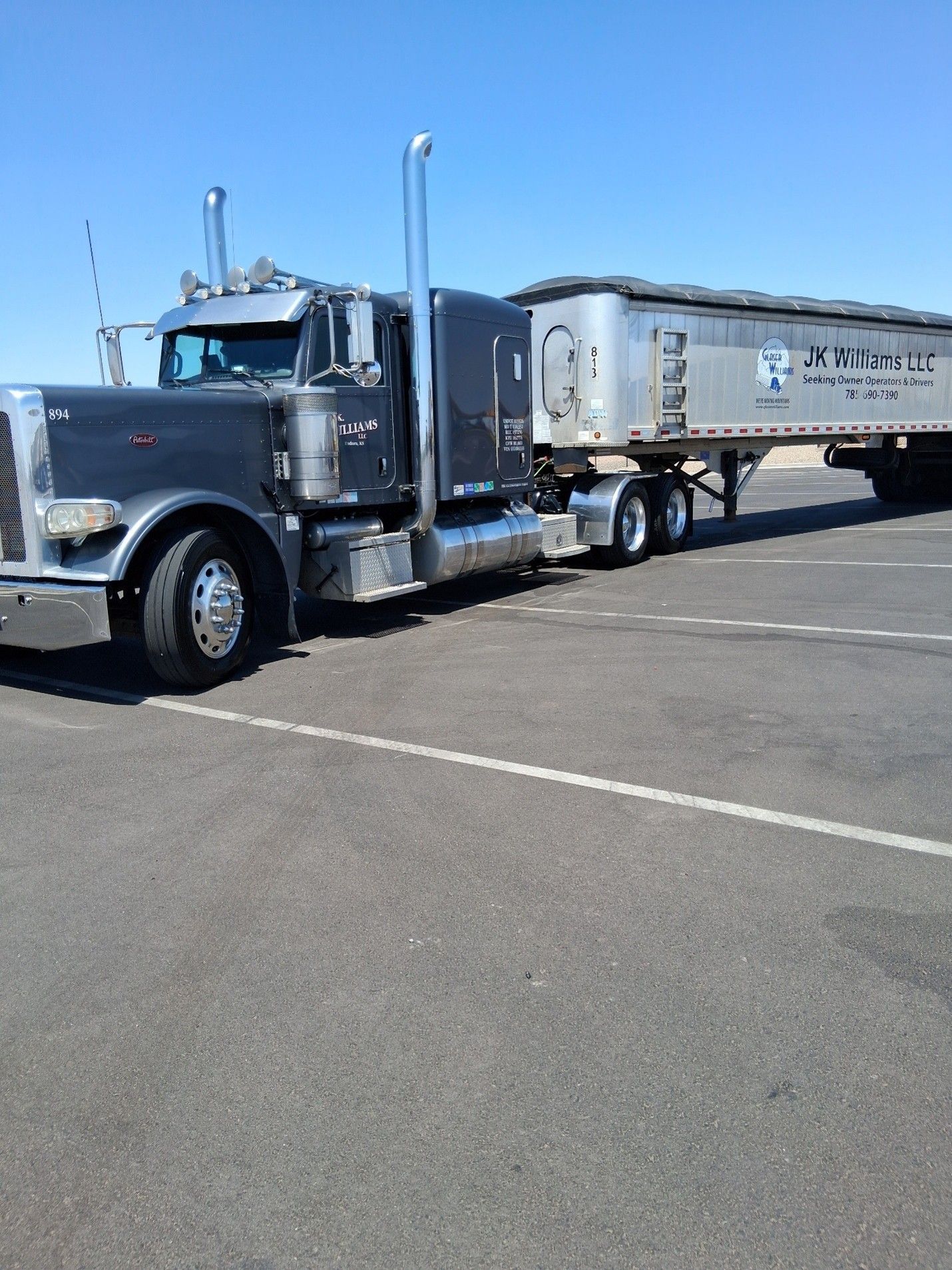 Silver semi-truck with flatbed trailer parked in a sunny lot under a clear blue sky