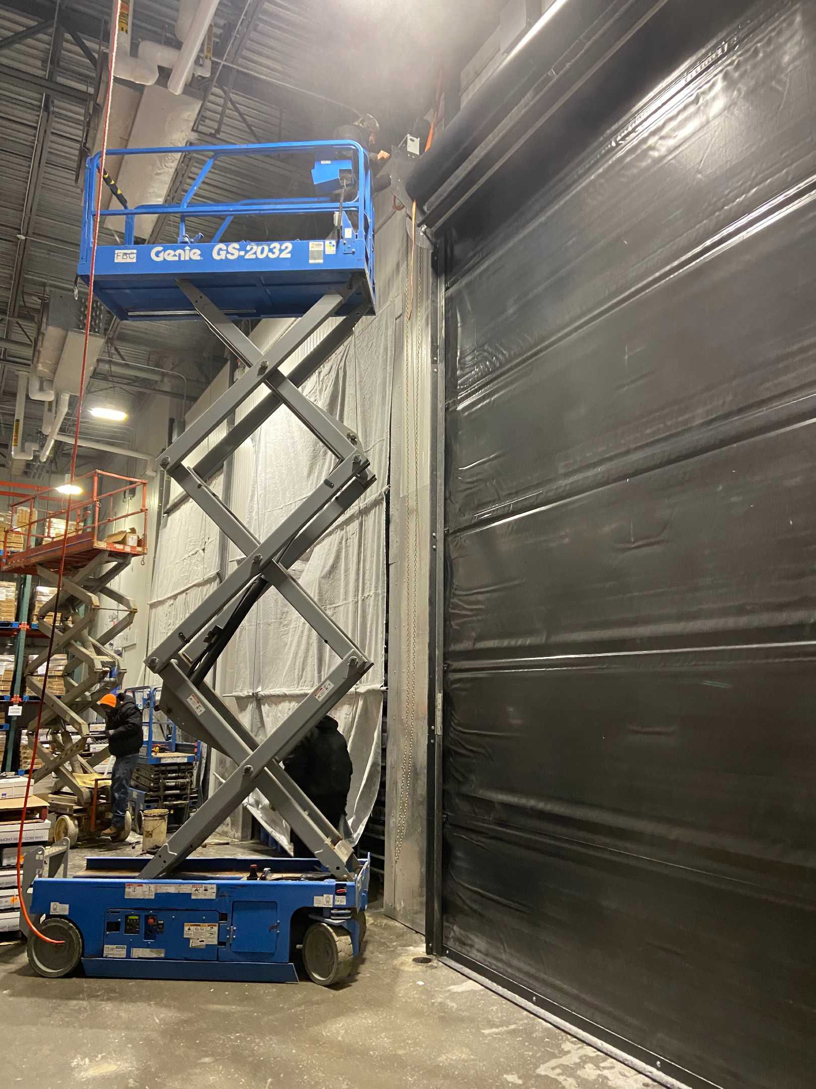 A blue scissor lift is sitting next to a black garage door in a warehouse.