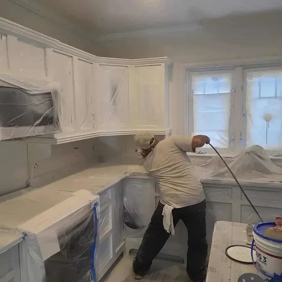 Person spray-painting white kitchen cabinets; surfaces are covered in plastic sheeting.