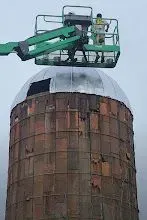 Workers in a lift installing a metal dome roof on a weathered, cylindrical silo.
