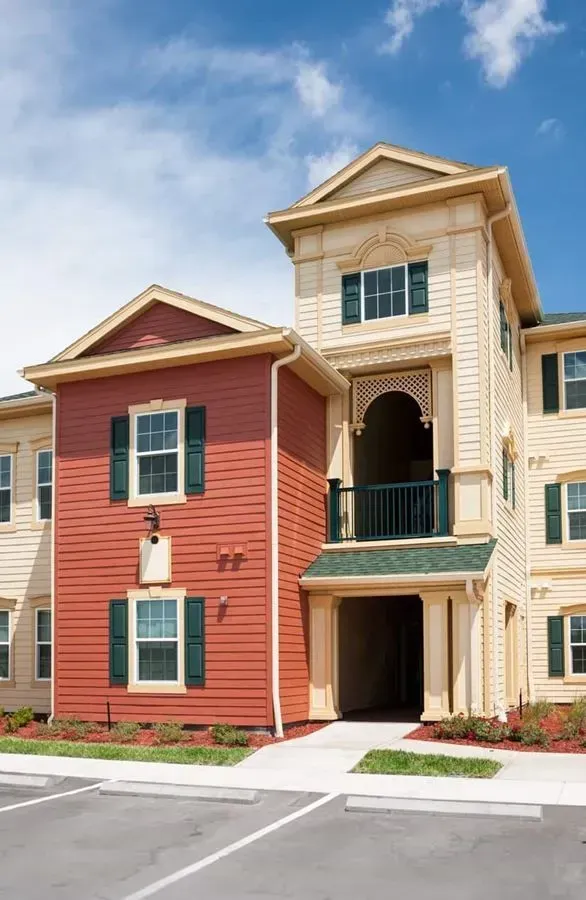 Apartment building exterior, red and tan siding, balcony, dark green shutters, blue sky.