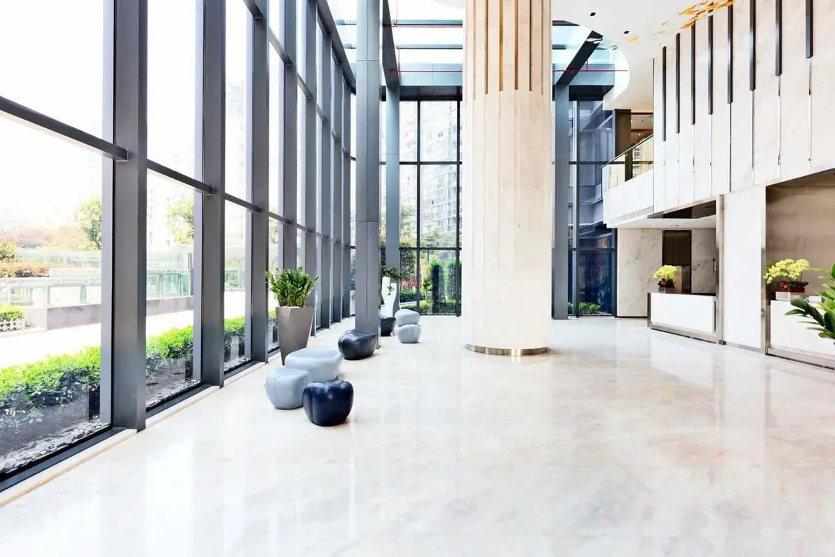 Modern lobby with large windows, light-colored flooring, and decorative seating. Tall pillars and greenery add to the space.