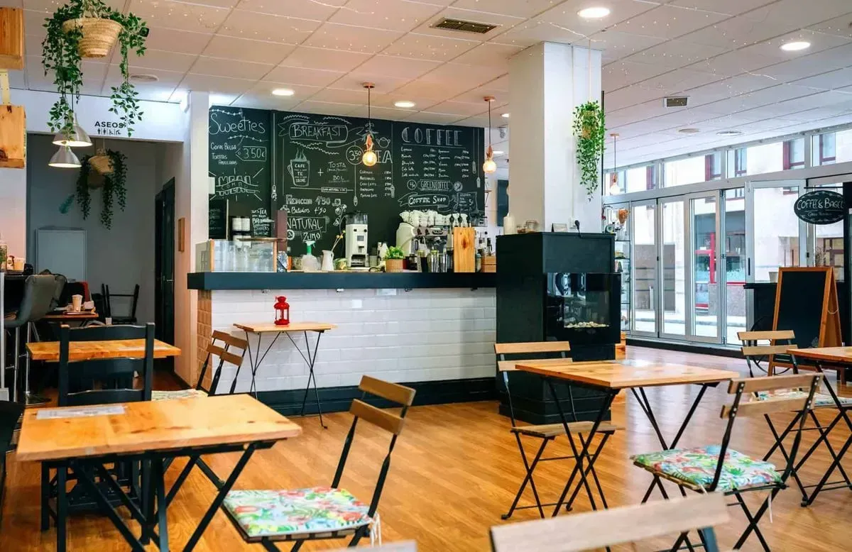 Coffee shop interior: wooden tables and chairs, chalkboard menu, white and black counter, plants, and large windows.