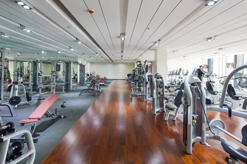 Gym interior with various exercise machines and equipment, wooden floor, and bright lighting.