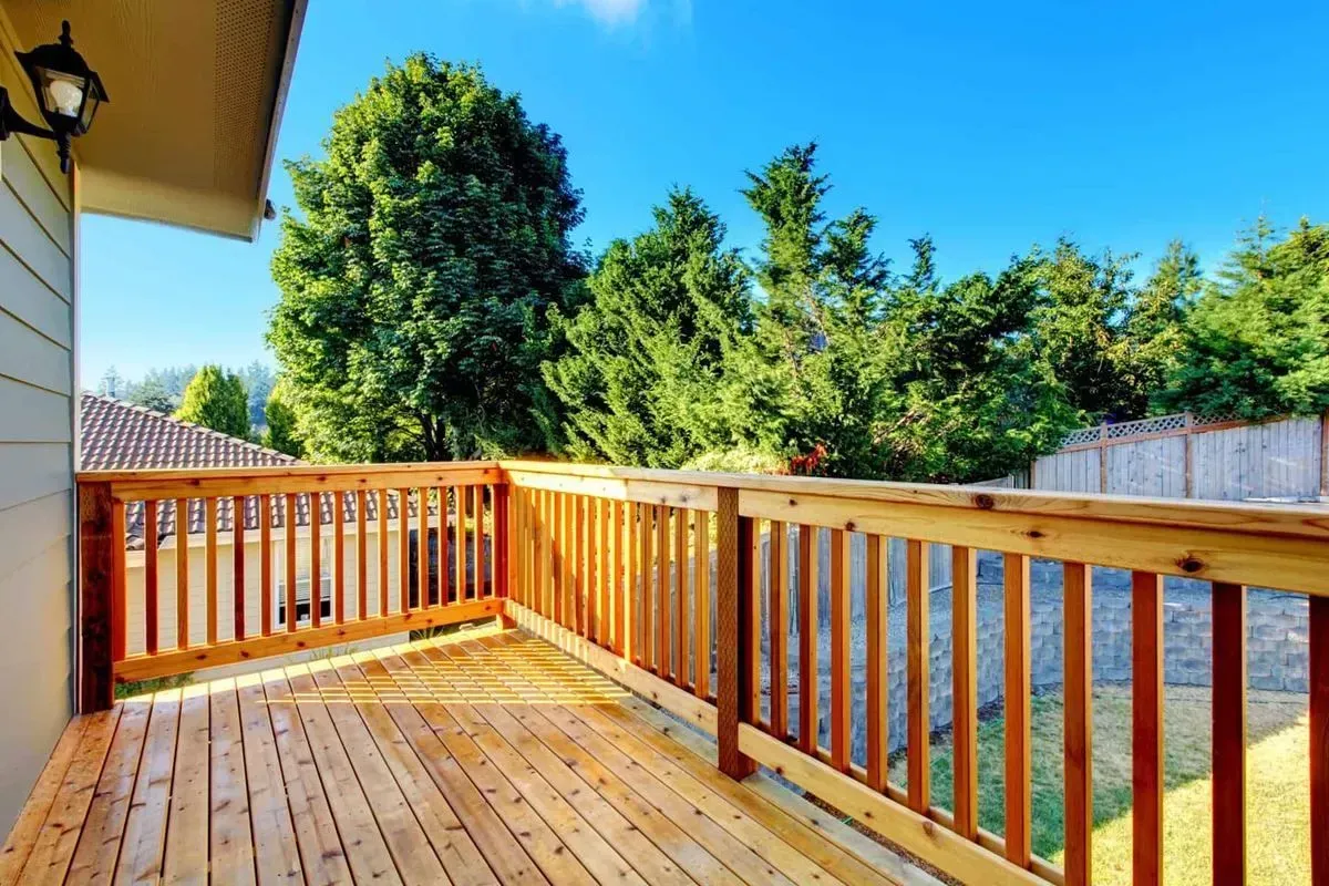Wooden deck with railings, overlooking a backyard with trees and a blue sky.