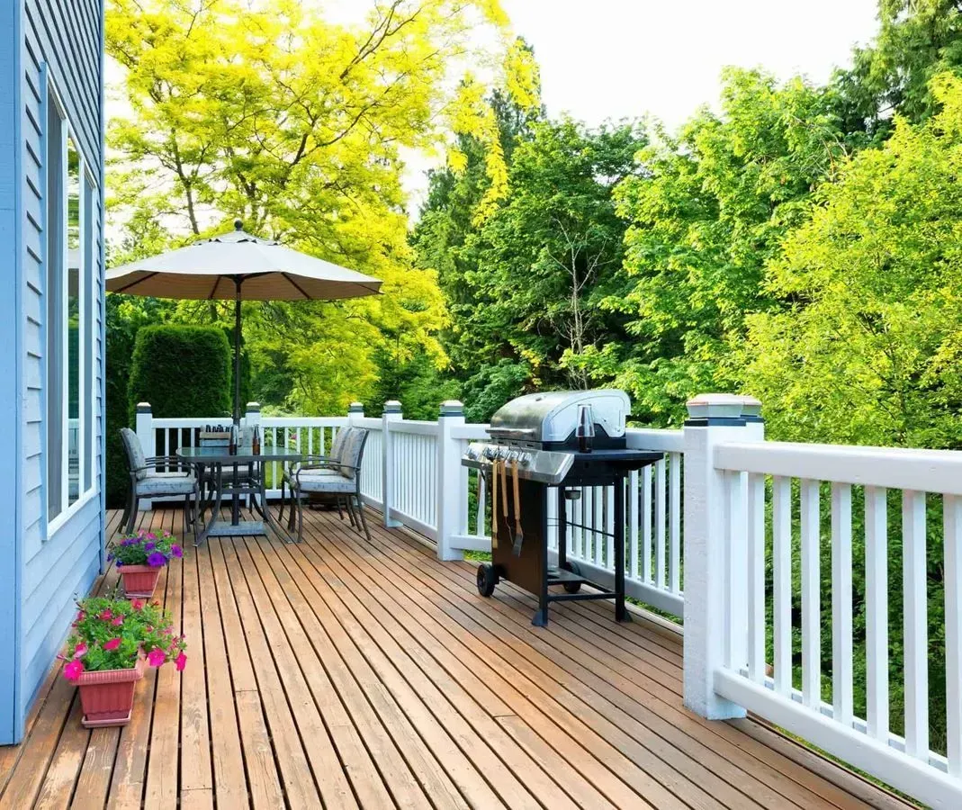 Wooden deck with white railing, patio furniture, and grill, overlooking lush green trees.