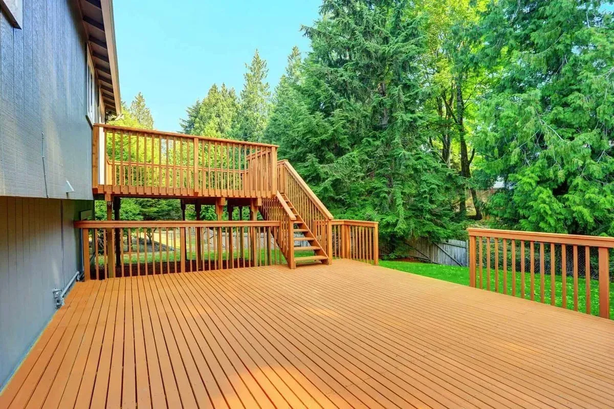Wooden deck with steps, surrounded by railings, adjacent to a house, green trees in background.