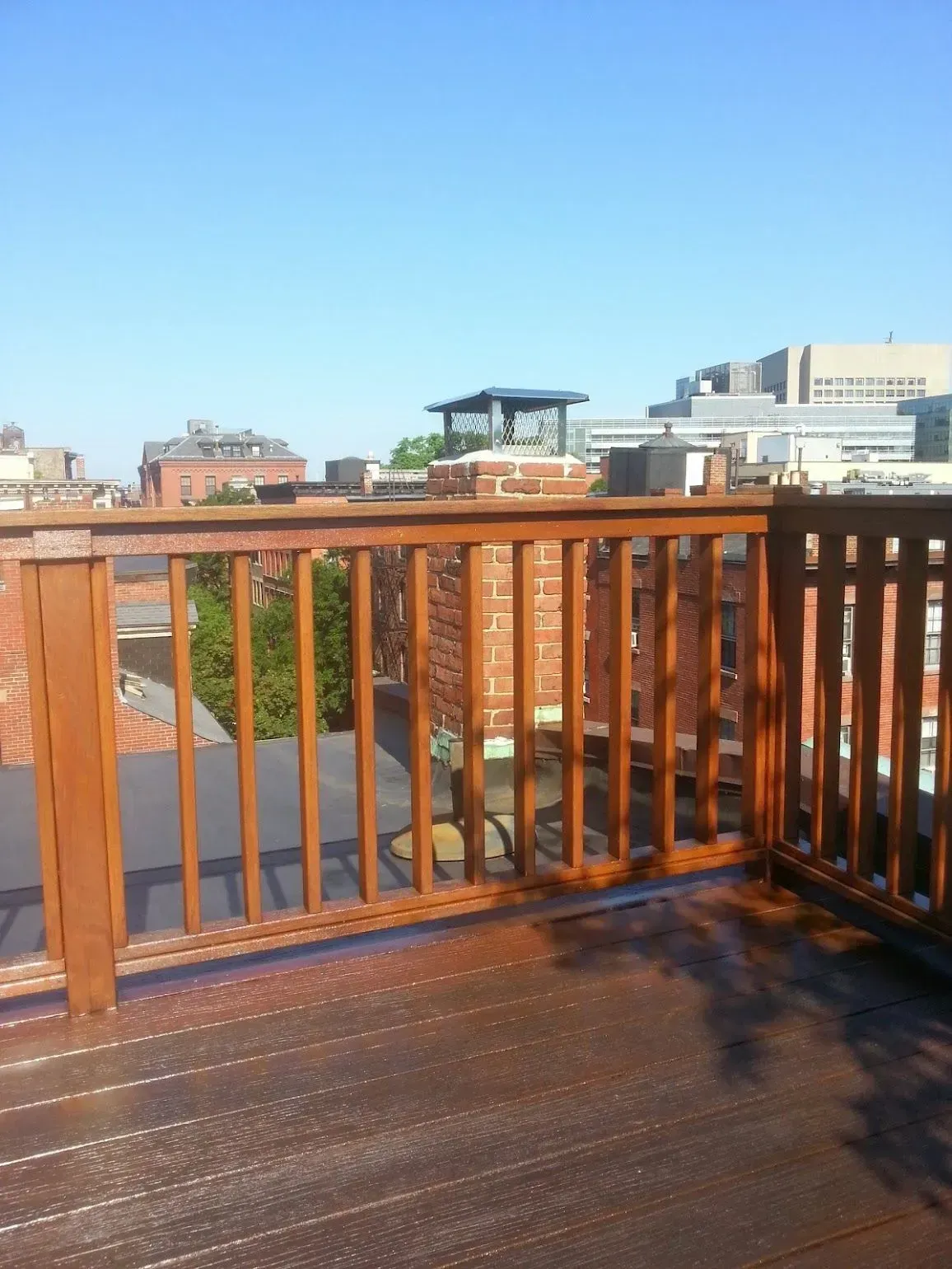 Wooden deck with railing, overlooking city buildings under a clear, blue sky.