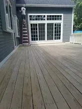 Wooden deck with person on a ladder painting a house's exterior wall. Gray siding, white windows.