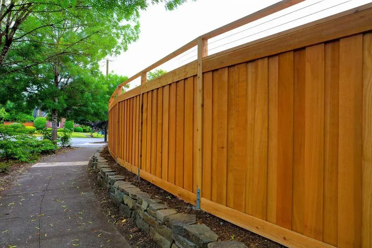 Wooden fence curving along a sidewalk, with greenery and trees visible in the background.