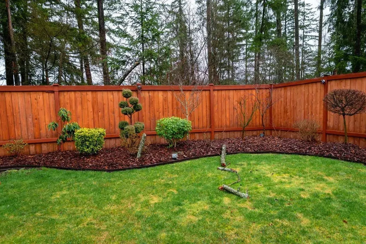 Lush green lawn with a flowerbed bordering a stained wooden fence; various trimmed shrubs.