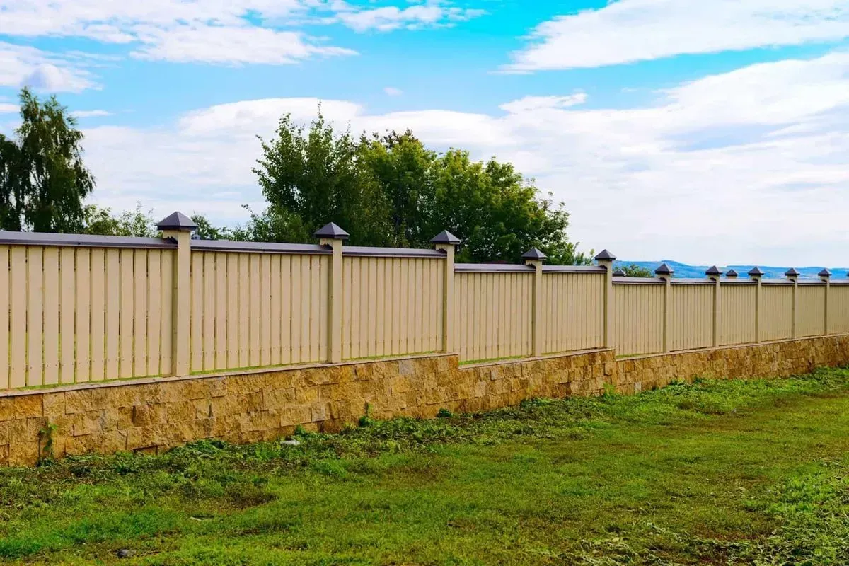 Tan fence with decorative posts atop a stone base, green grass in front, blue sky with clouds.