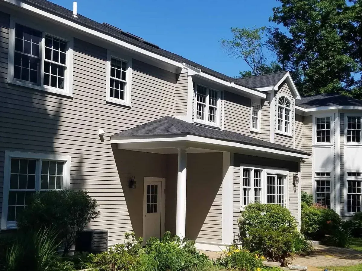 Two-story house with gray siding, white trim, and a dark roof under a blue sky, surrounded by greenery.