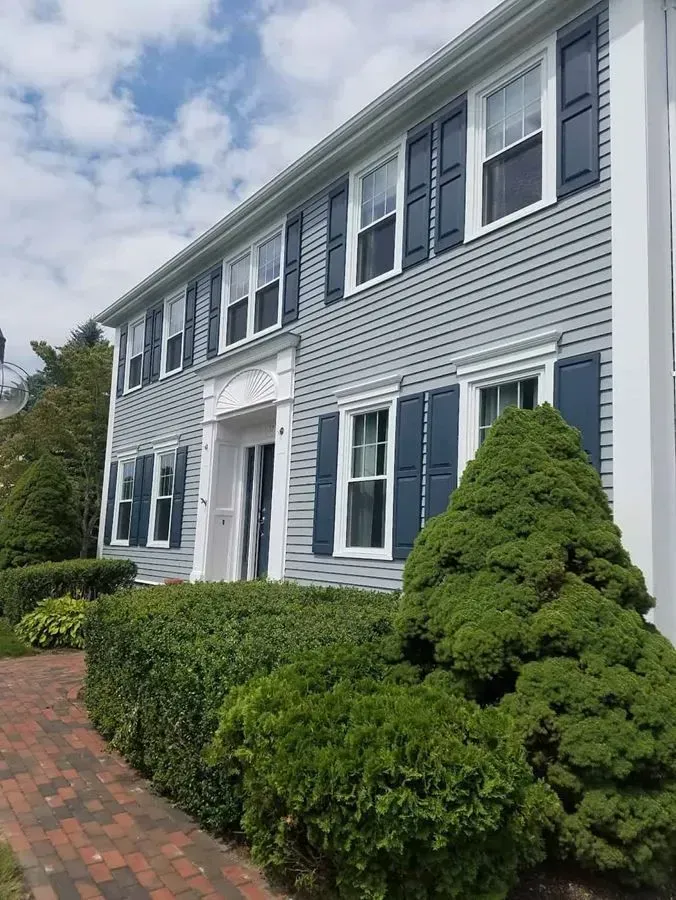 Two-story, light gray house with dark blue shutters and white trim, surrounded by green bushes, and a brick pathway.
