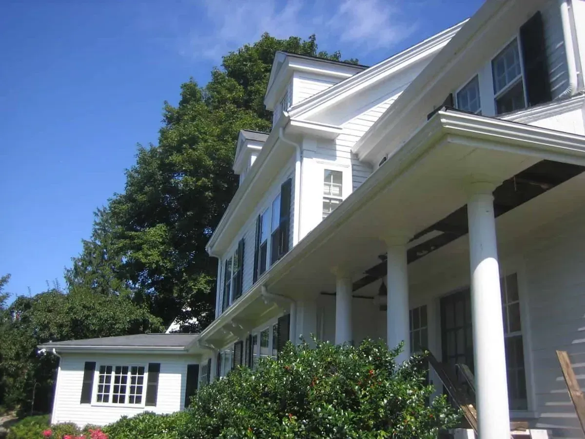 White house with dark shutters, white columns, blue sky and green trees.