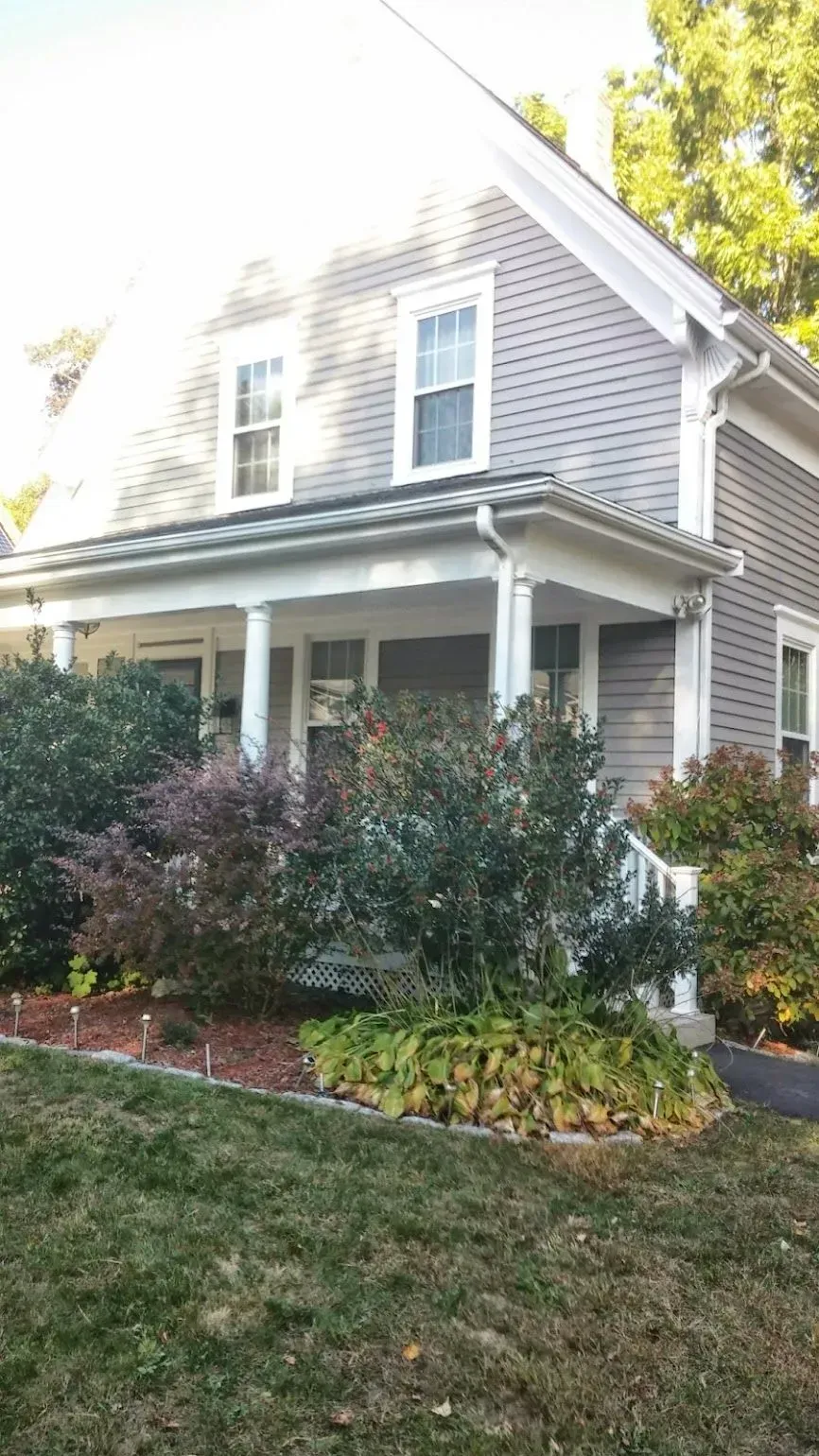 Gray shingled house with porch, shrubs, and green lawn.