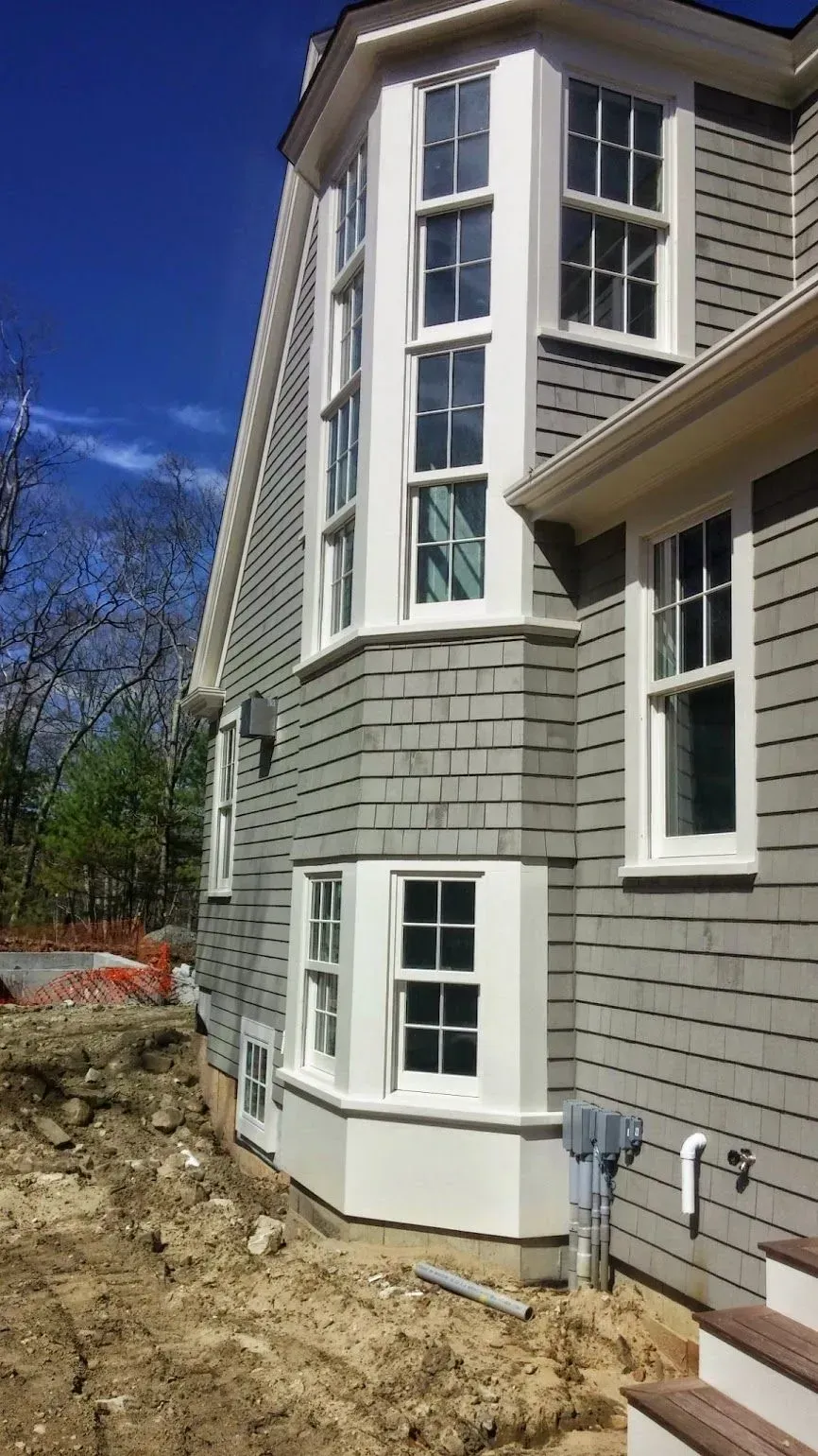 Gray house with white-framed windows, bay window, and gray shingles. Construction site in the foreground.