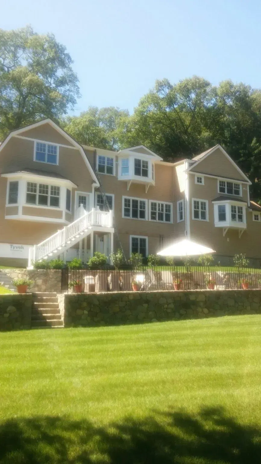 Beige house with a white staircase, a stone wall, and a large lawn.