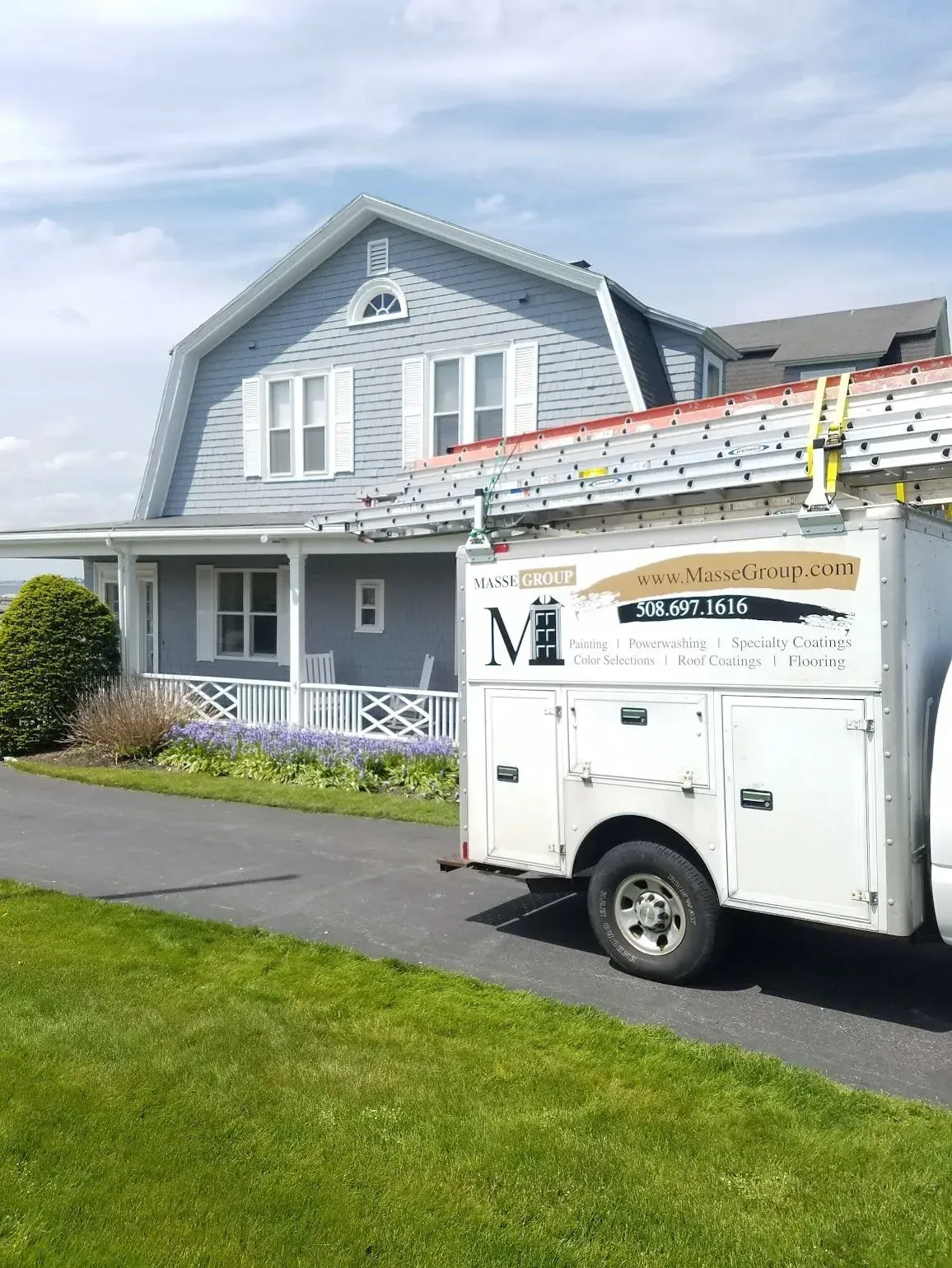 A white work truck with ladder in front of a gray house. Green grass and blue sky are visible.