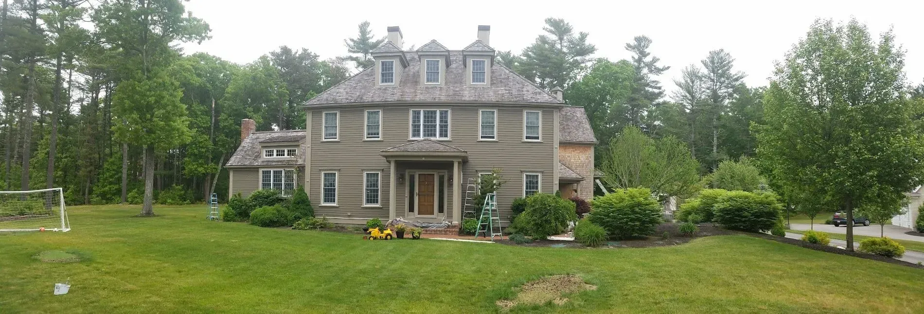 Large two-story house with green lawn, trees, and sky. The house is tan with many windows.