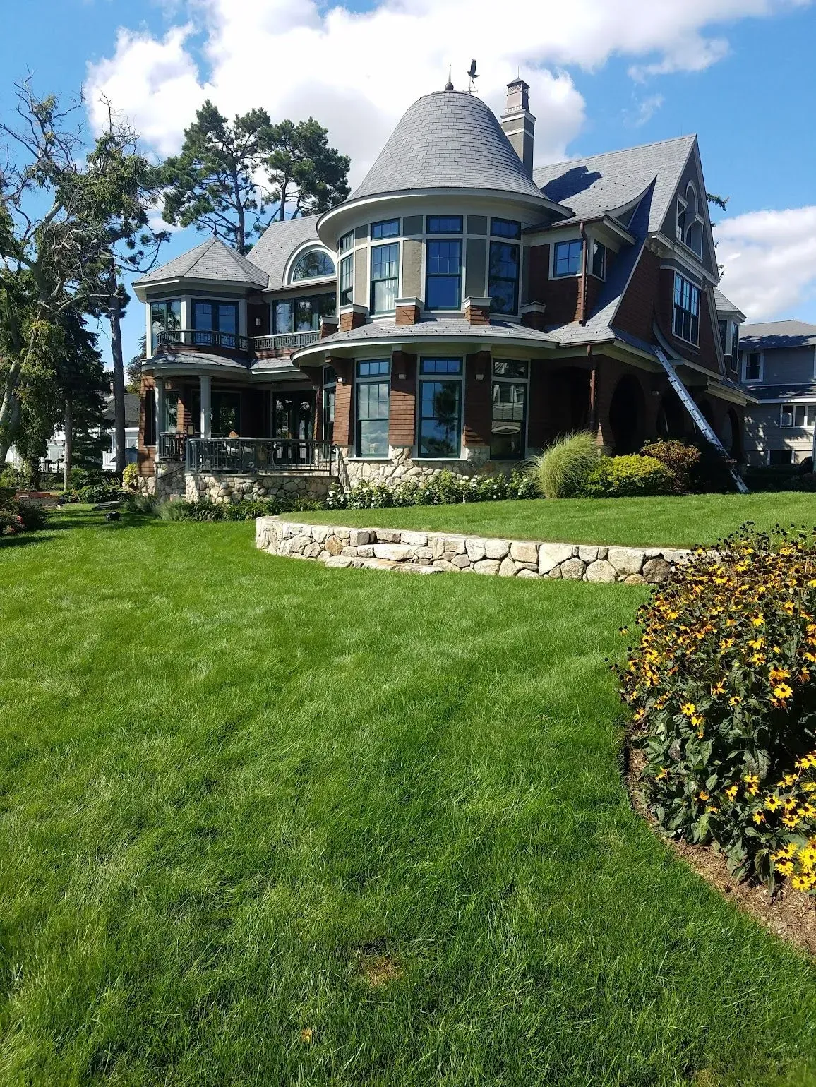 Large brown house with grey roof, multiple windows, green lawn, and blue sky.