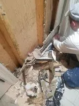 Man repairing damaged wall near a window; debris on the floor, wood framing visible.