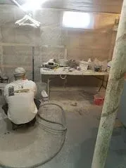 Person in white shirt working in a cluttered basement. A table with items sits in the center. Gray walls.