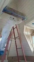 Person in protective suit spraying a ceiling from a ladder in a building under construction.