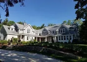 Large gray house with many windows and gabled roof, surrounded by trees and a blue sky.