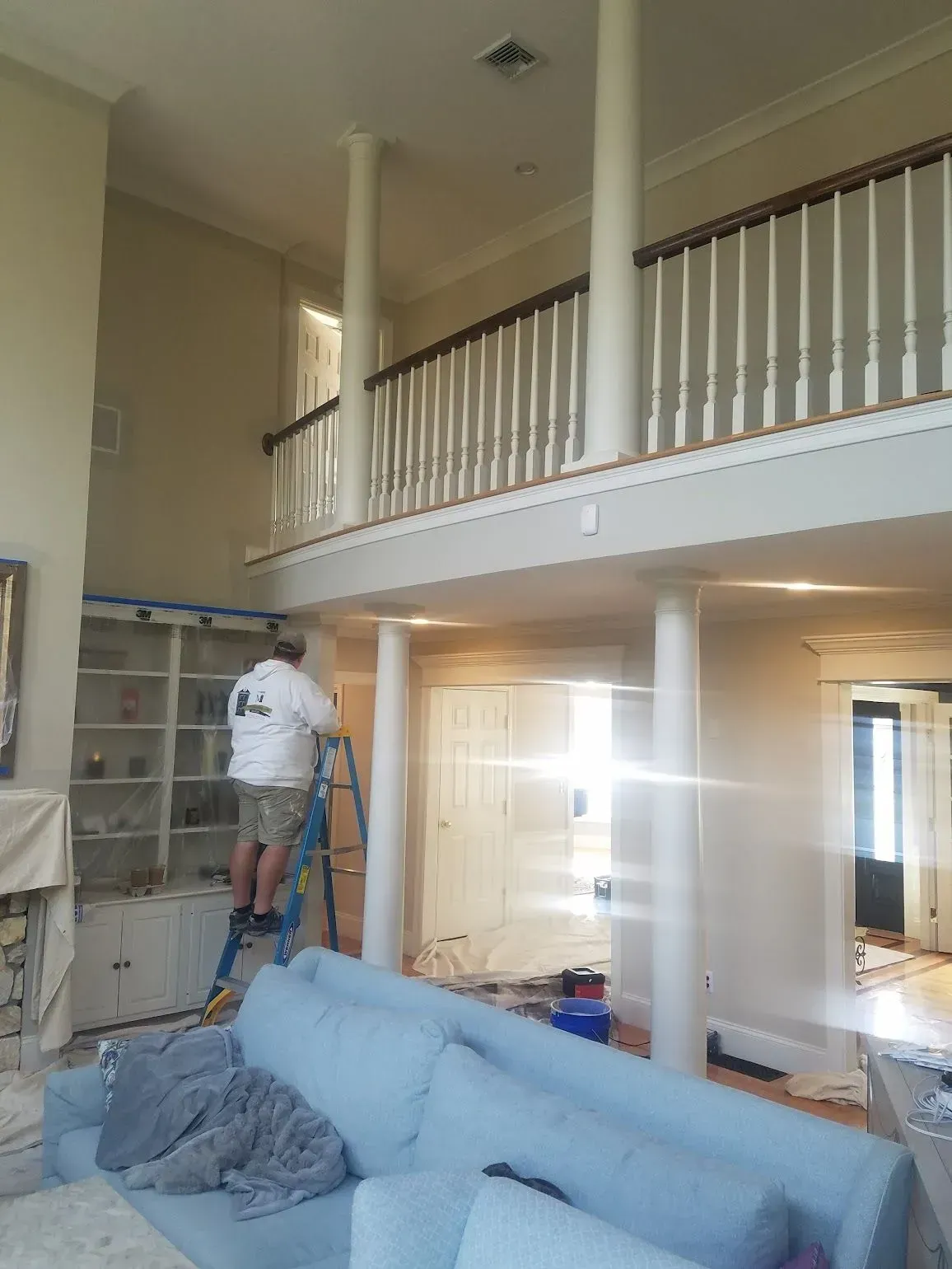 Person painting tall interior wall from a ladder, with columns, a balcony, and a blue sofa.