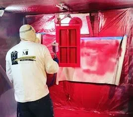 Person spraying red paint on a window frame inside a red-lined spray booth.