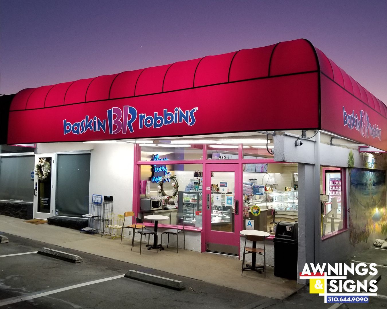 Baskin-Robbins store exterior with pink awning, pink door, and small tables. The sky is dusky.