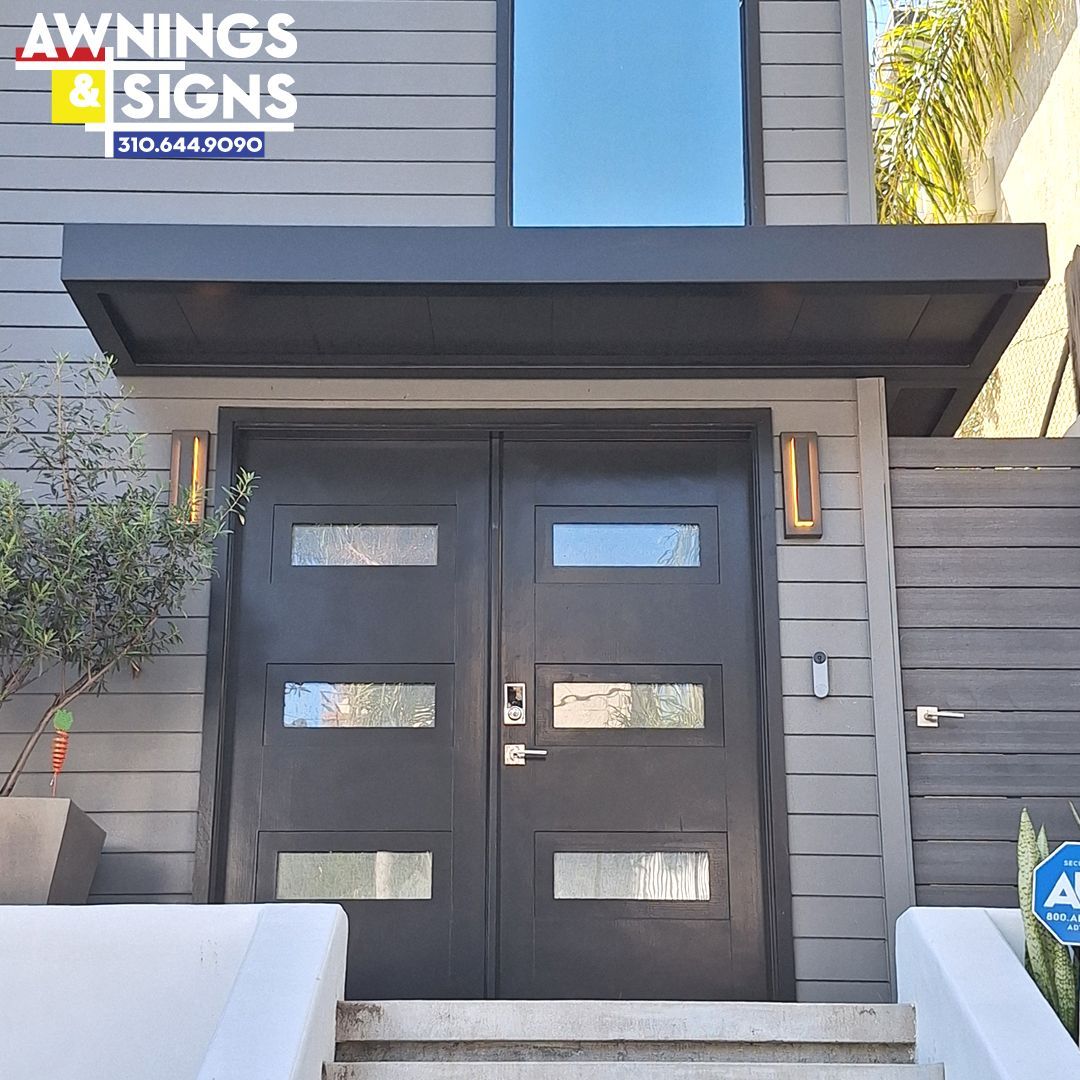 Gray double doors with rectangular glass panes and a dark awning over a gray building.