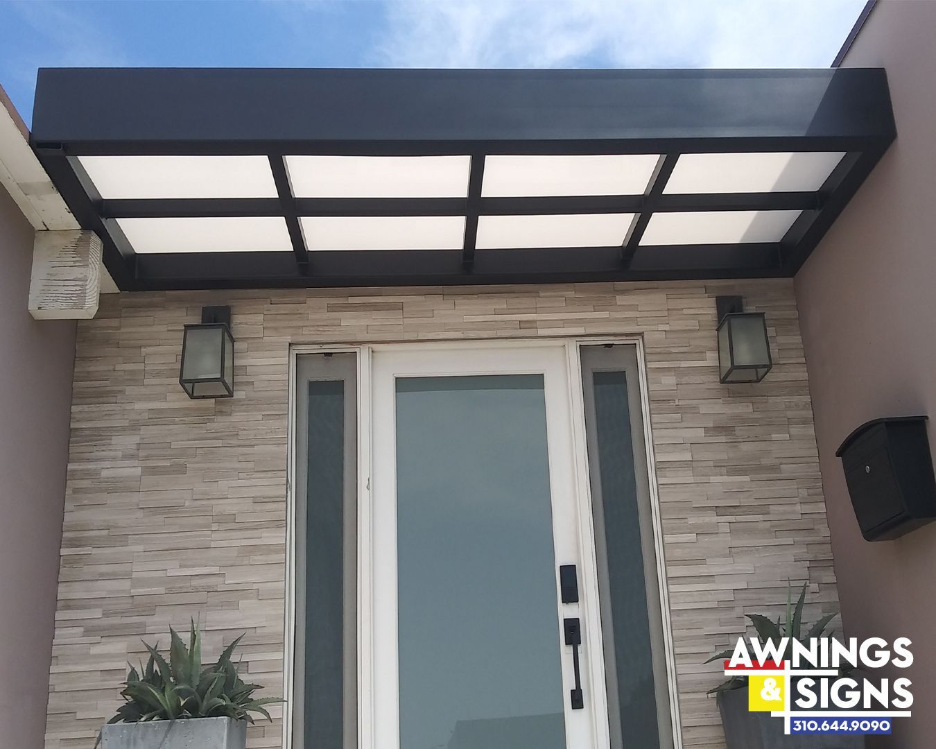 Black awning above a front door with sidelights, brick-like siding, and light fixtures.