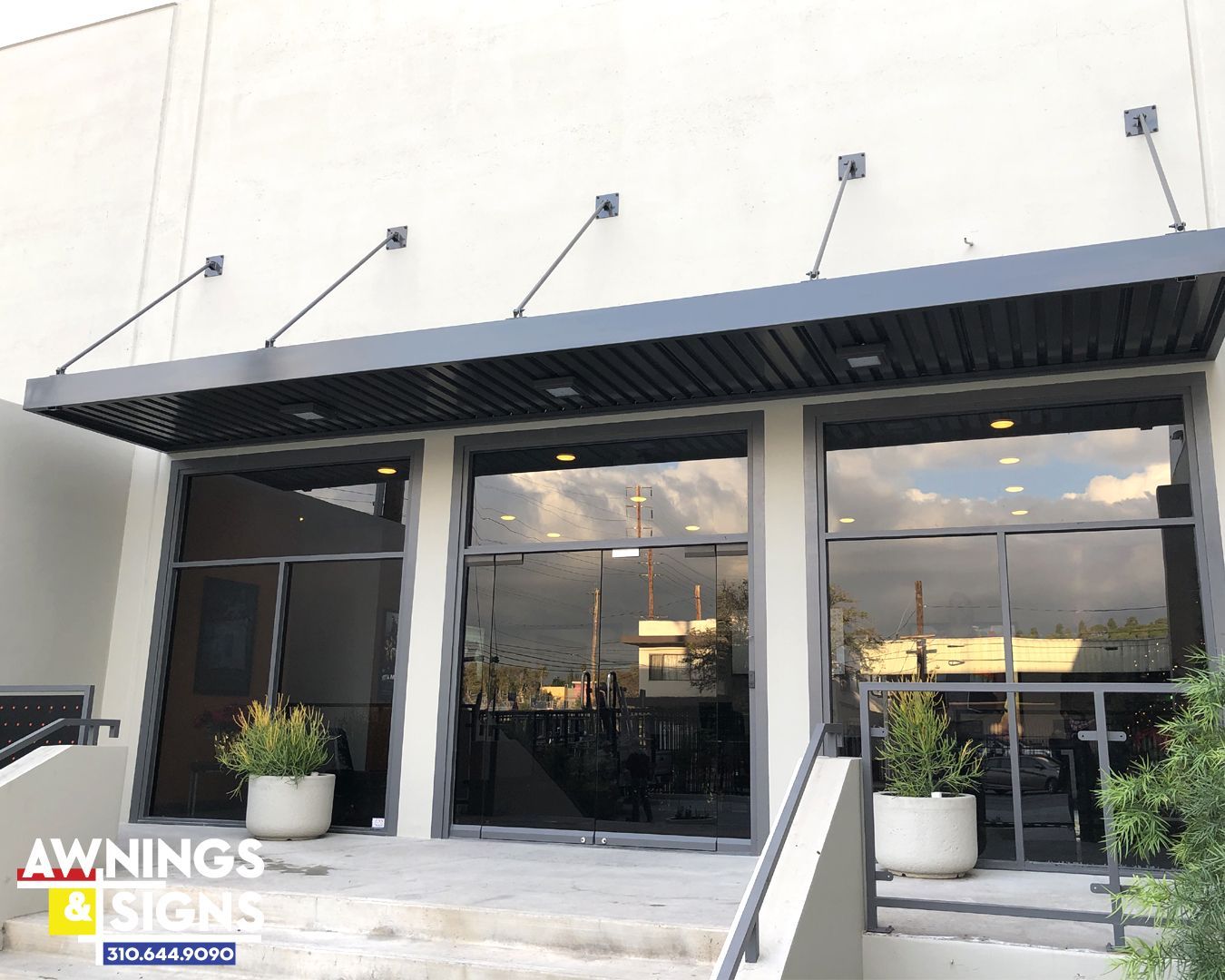 Gray building entrance with black awnings, glass windows, and potted plants.