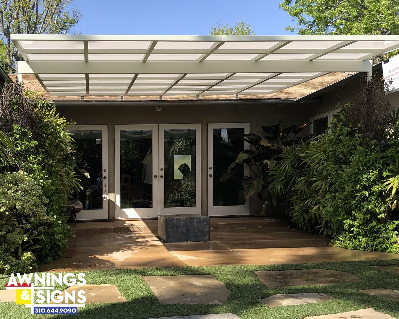 White patio cover over French doors, surrounded by green plants, in a yard.