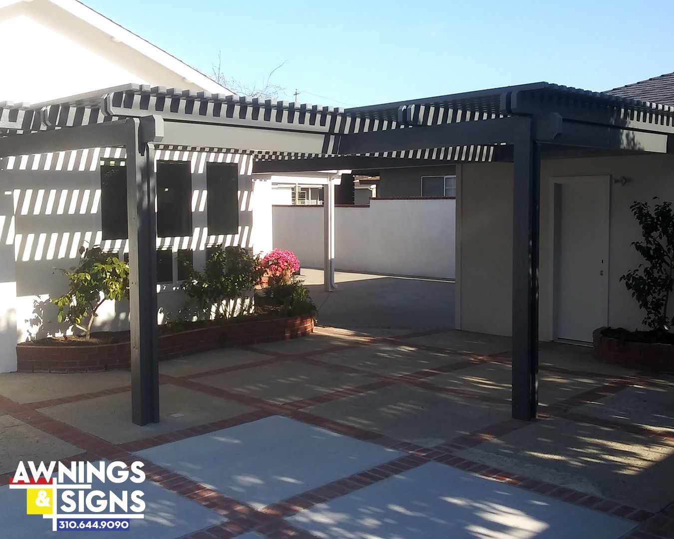 Gray pergola over a paved patio with red brick borders next to a white building.
