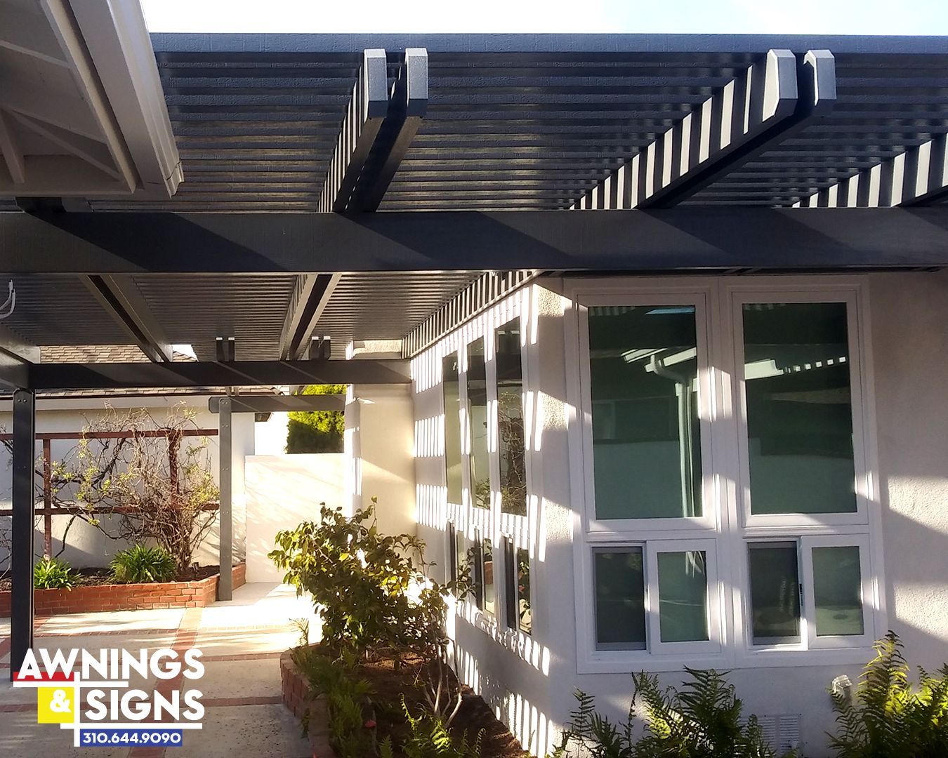 Pergola over windows with a walkway. White walls, dark beams, plants, bright sun.