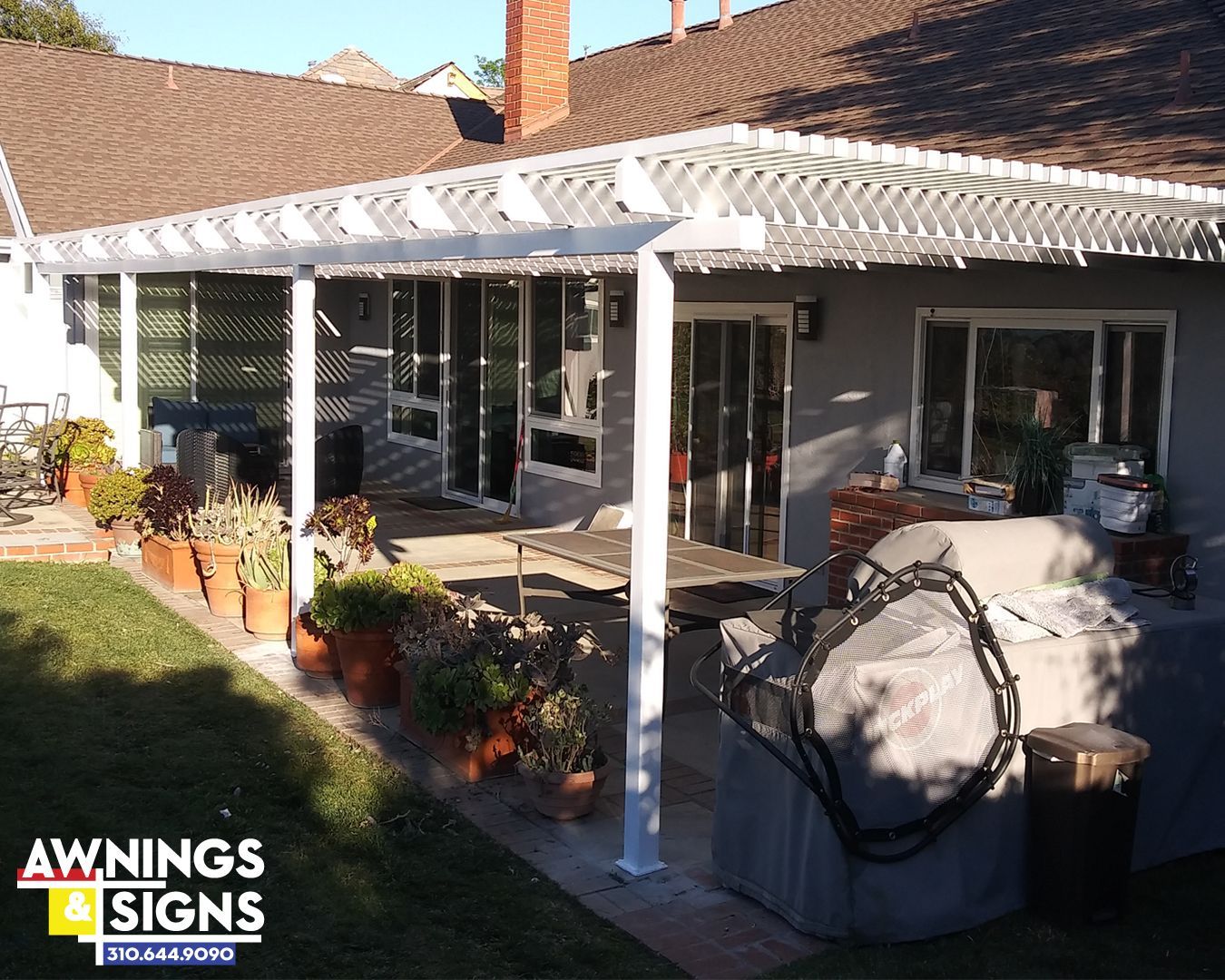 White pergola over a patio with potted plants, sliding glass doors, and a grill covered in a tarp.
