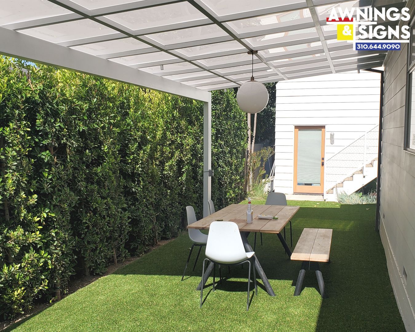 Outdoor dining area with pergola, table, chairs, and artificial grass. Green hedge wall.