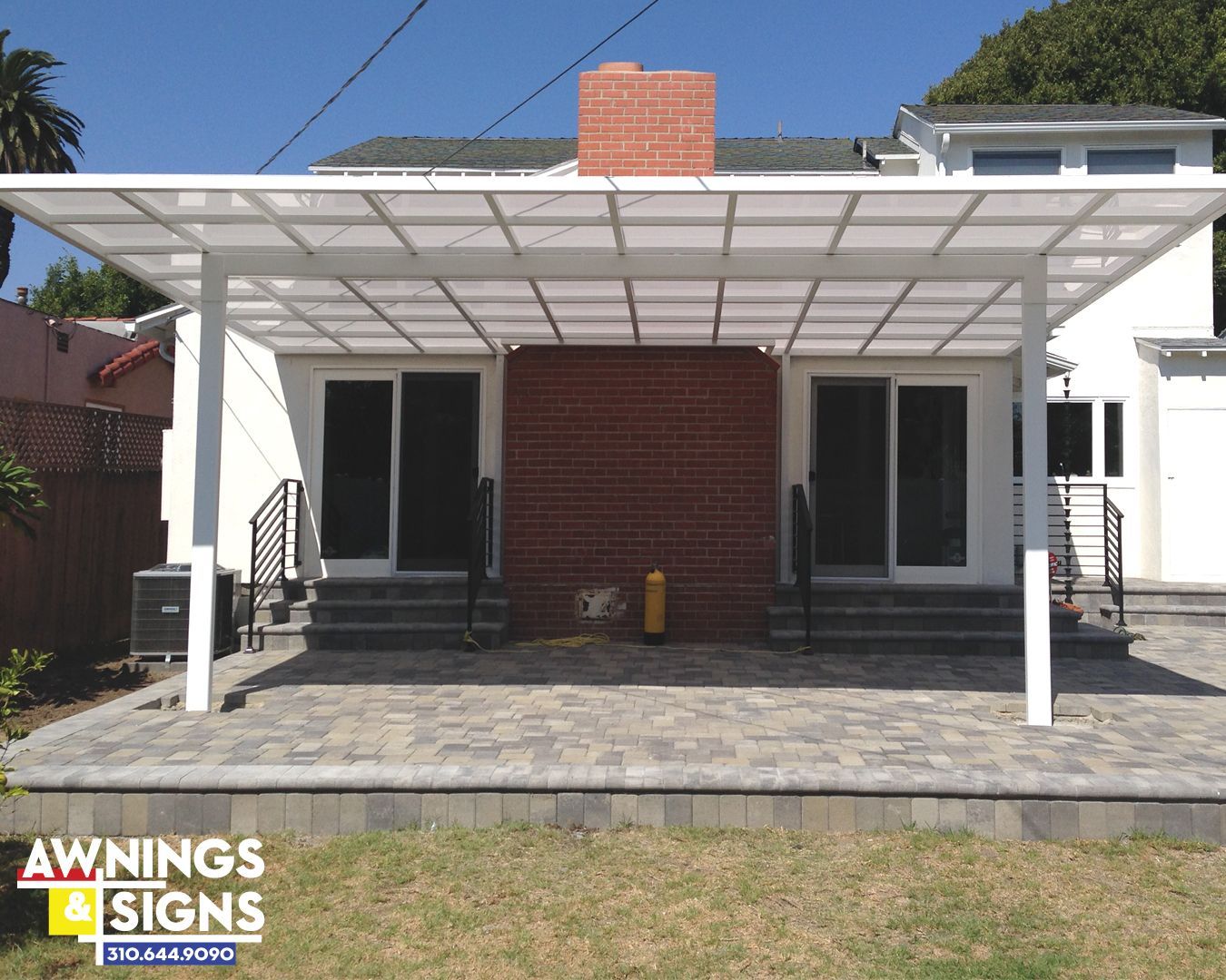 White patio cover over brick wall with two sliding doors, on stone patio with grass.