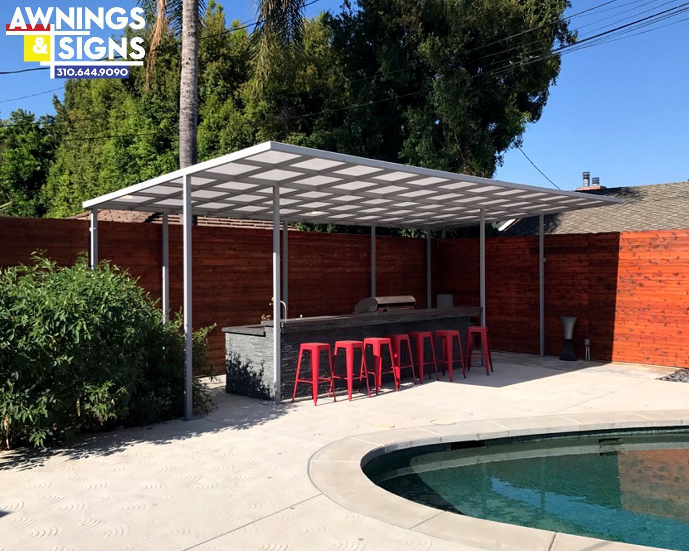 Outdoor kitchen with red stools, beneath a canopy near a pool.