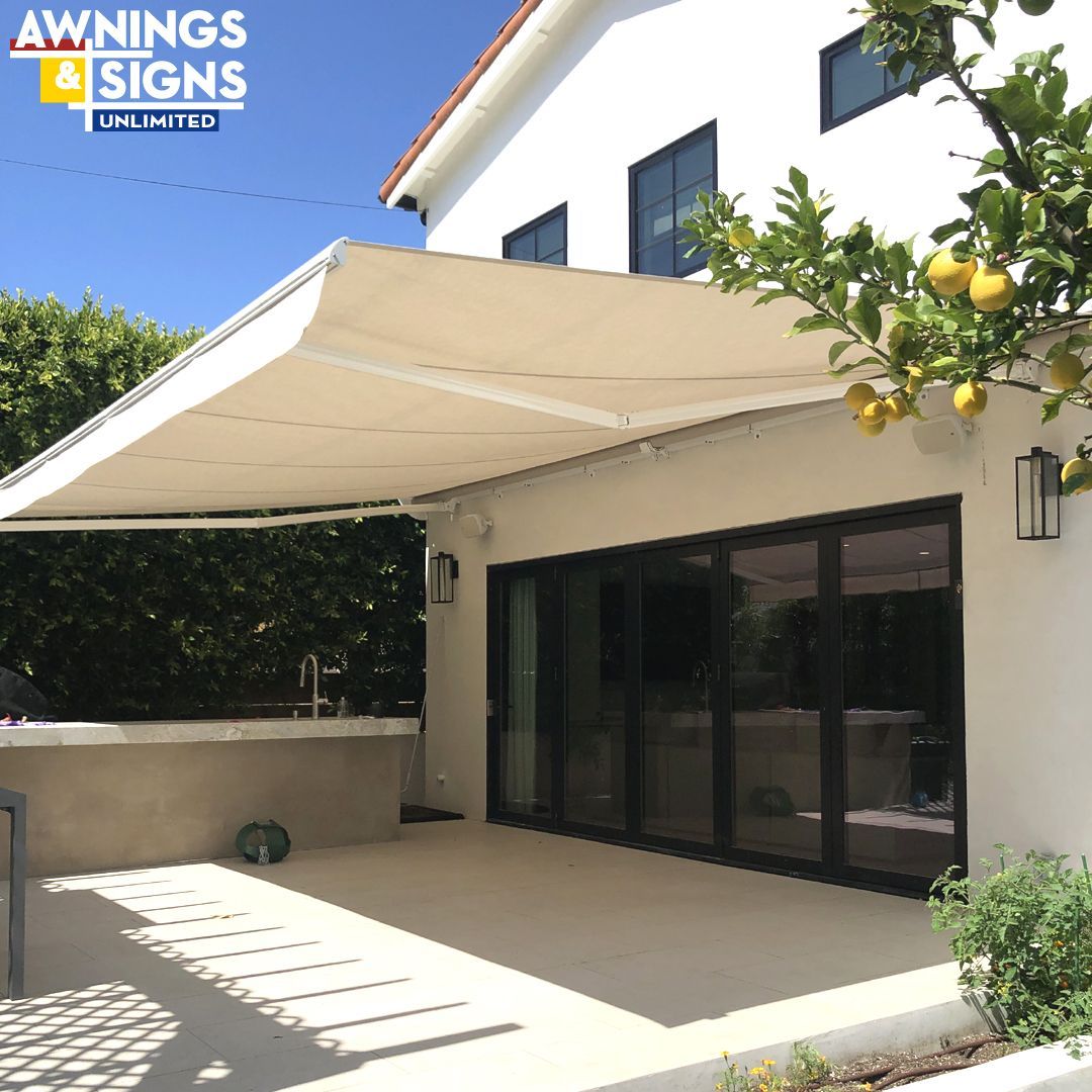 Tan retractable awning over a patio, next to a white house. Green foliage and glass doors are visible.