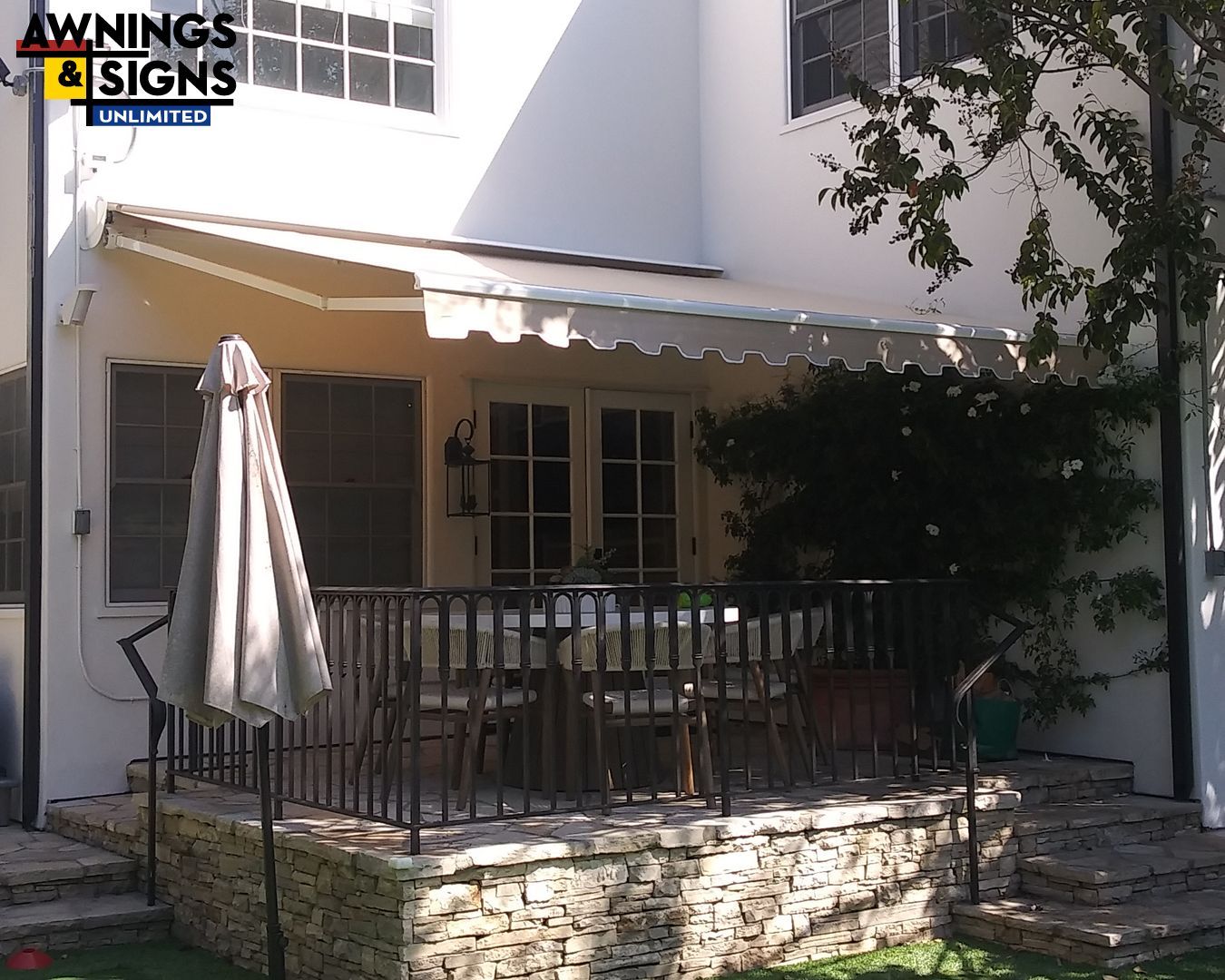 Beige awning over a patio with a table, chairs, and a railing. White house with a stone base.