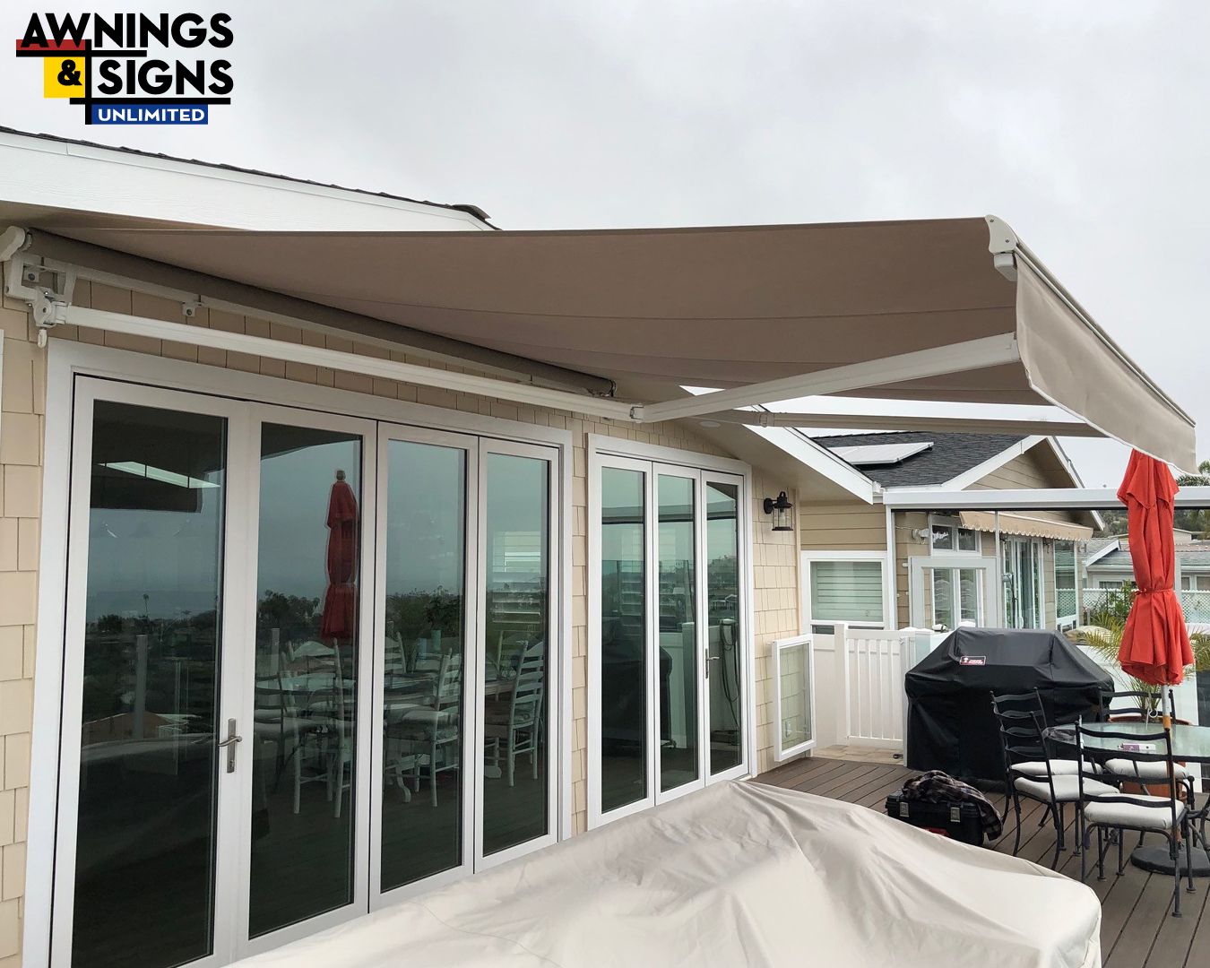 Beige retractable awning over glass doors on a deck with a grill and ocean view.