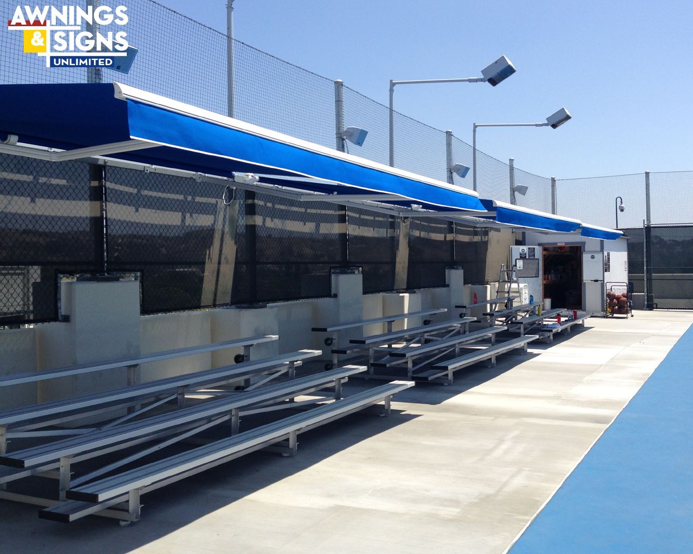 Bleachers under a blue awning at a tennis court, next to a black fence, under blue sky.