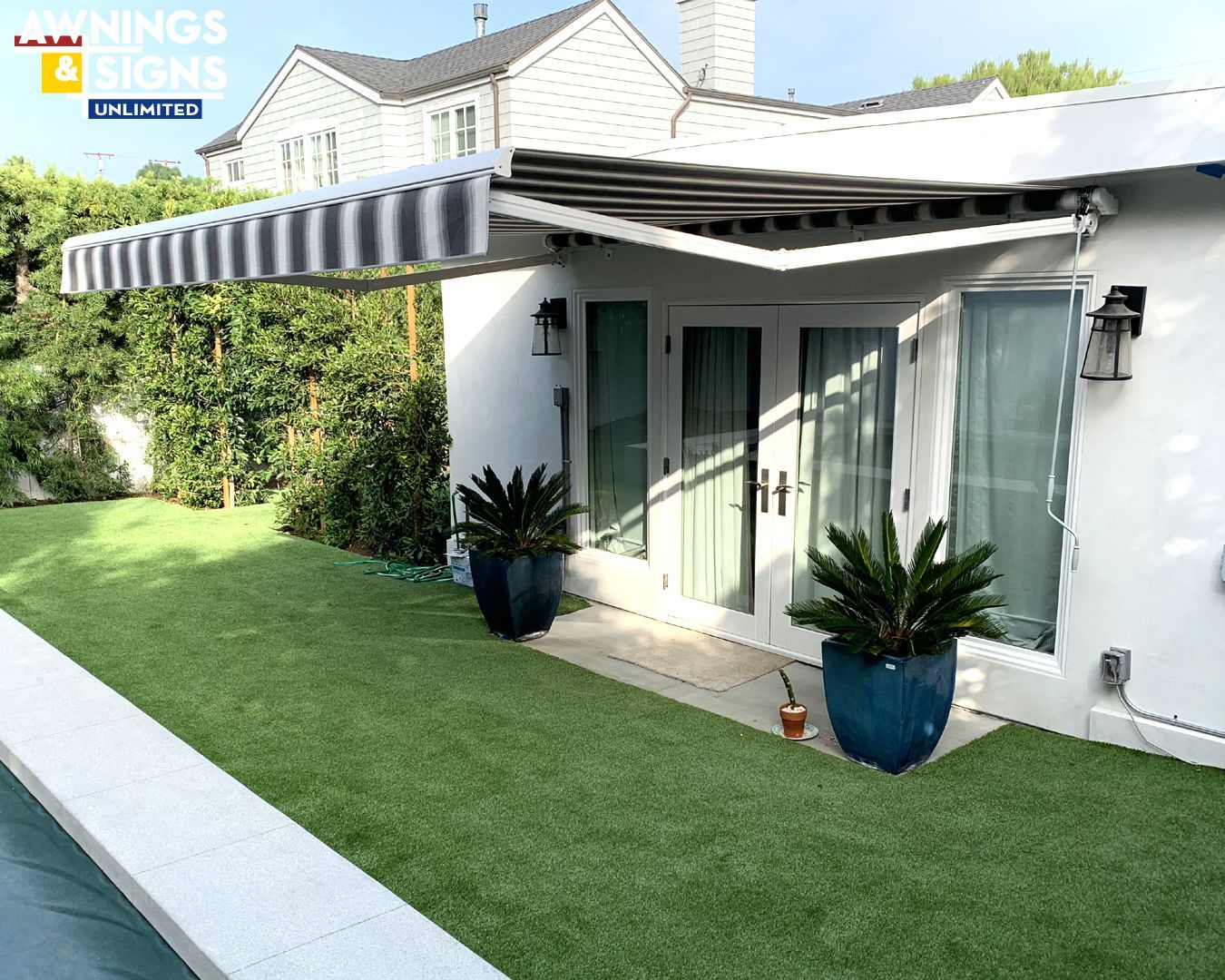 Exterior view: striped awning over white building's patio doors, flanked by potted plants on green lawn.