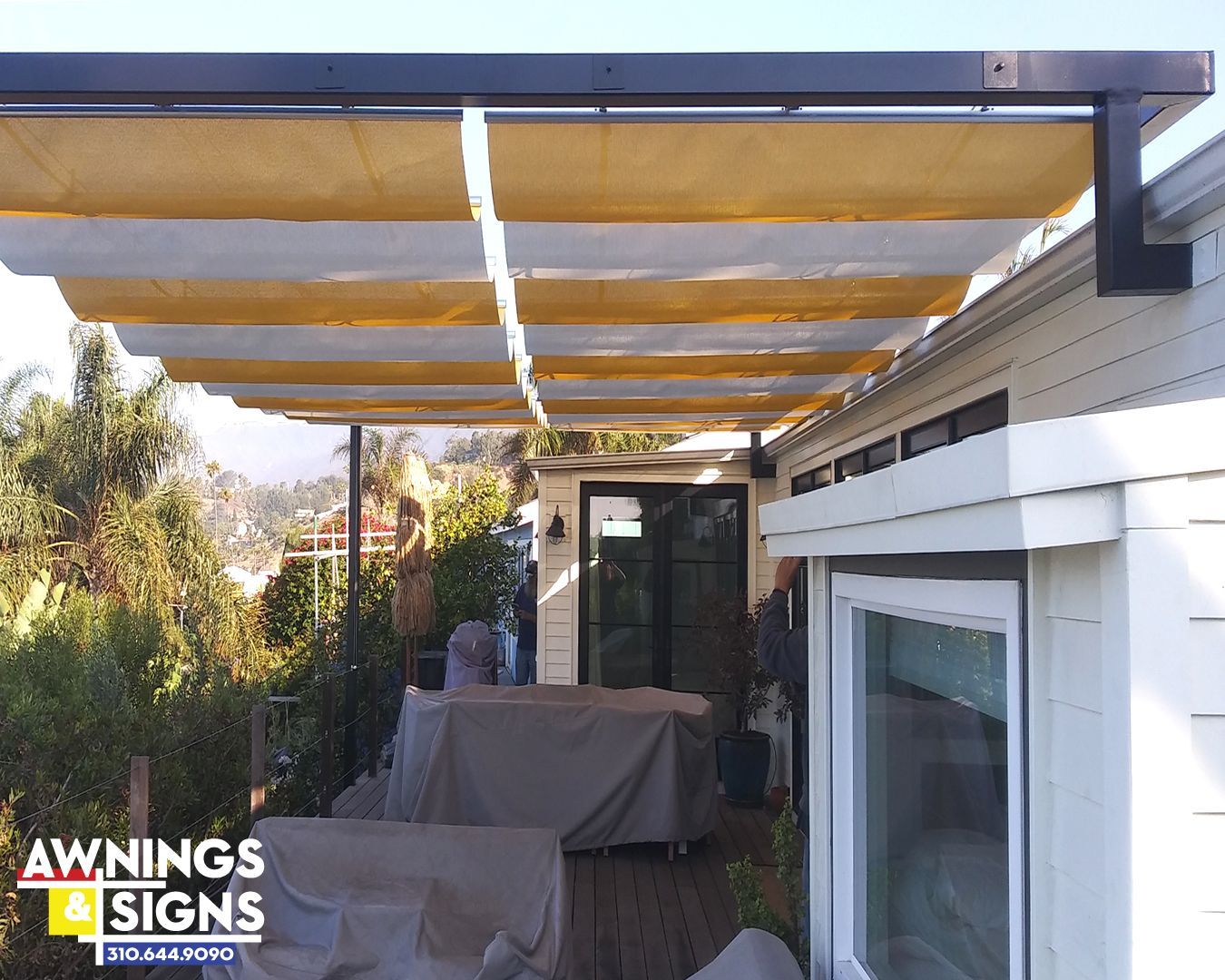 Pergola with cream and black shades, attached to a white house overlooking a deck and greenery.