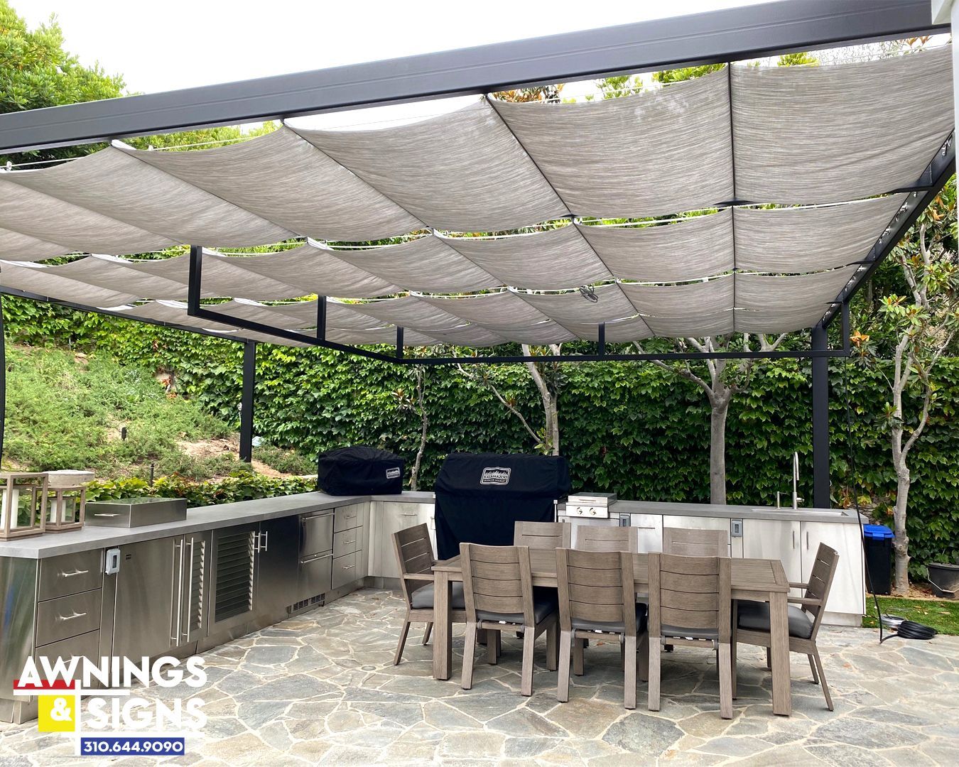 Outdoor kitchen with gray shade canopy, dining table, stainless steel cabinets, and grill.
